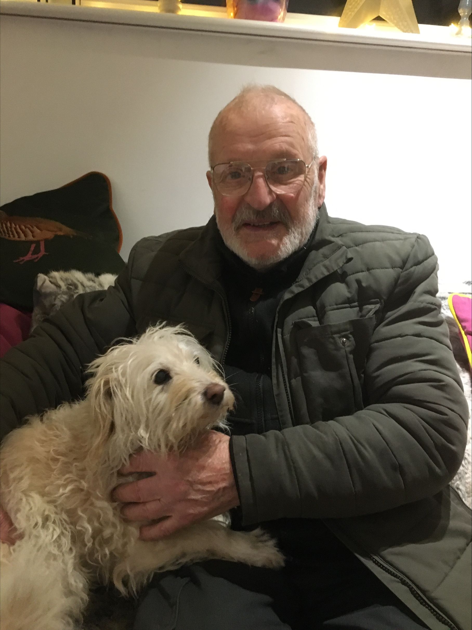 Elderly man with glasses and a white beard, smiling while wearing a padded jacket and holding a small fluffy white dog on his lap.