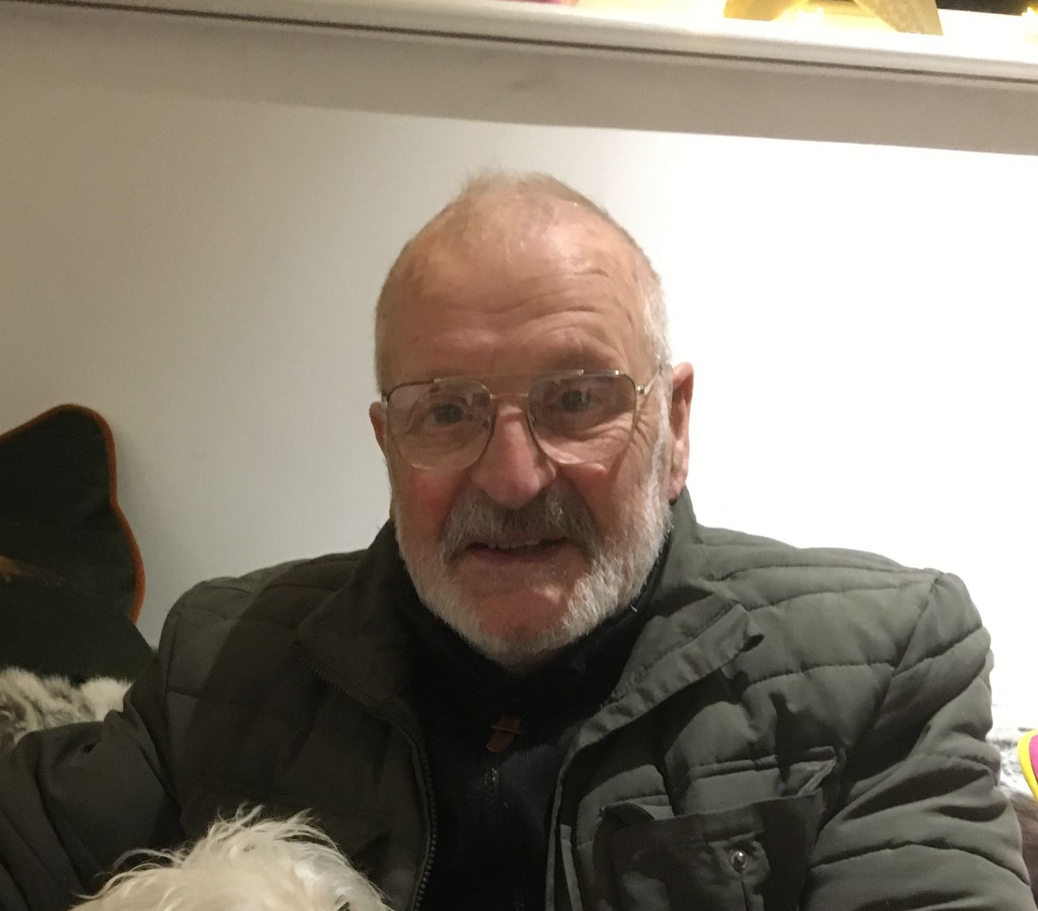Elderly man with glasses and a white beard, smiling while wearing a padded jacket and holding a small fluffy white dog on his lap.