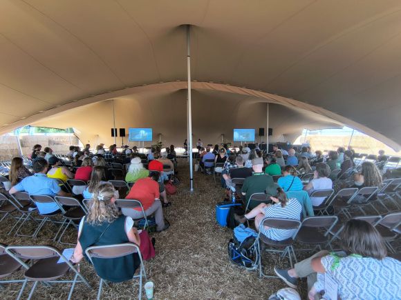 The interior of a tent at a festival. showing the backs of a crowd seated listening to a presentation. There is straw on the floor.