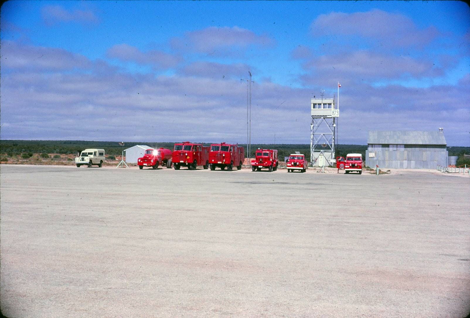 Bright red fire engines and land rovers sit on the runway next to a control tower on Maralinga airfield.