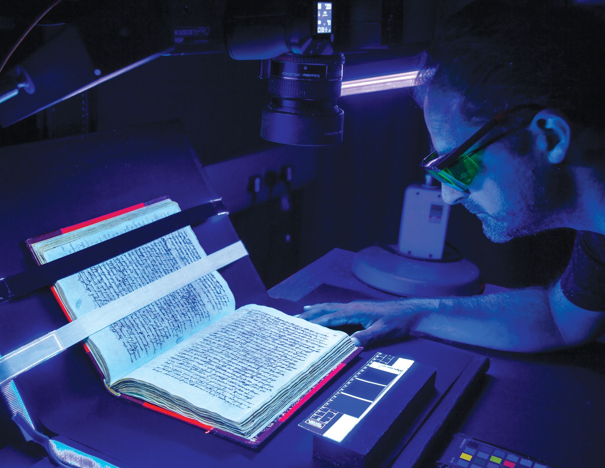 A man wears yellow glasses and looks closely at a book during multi-spectral imaging at the British Library.