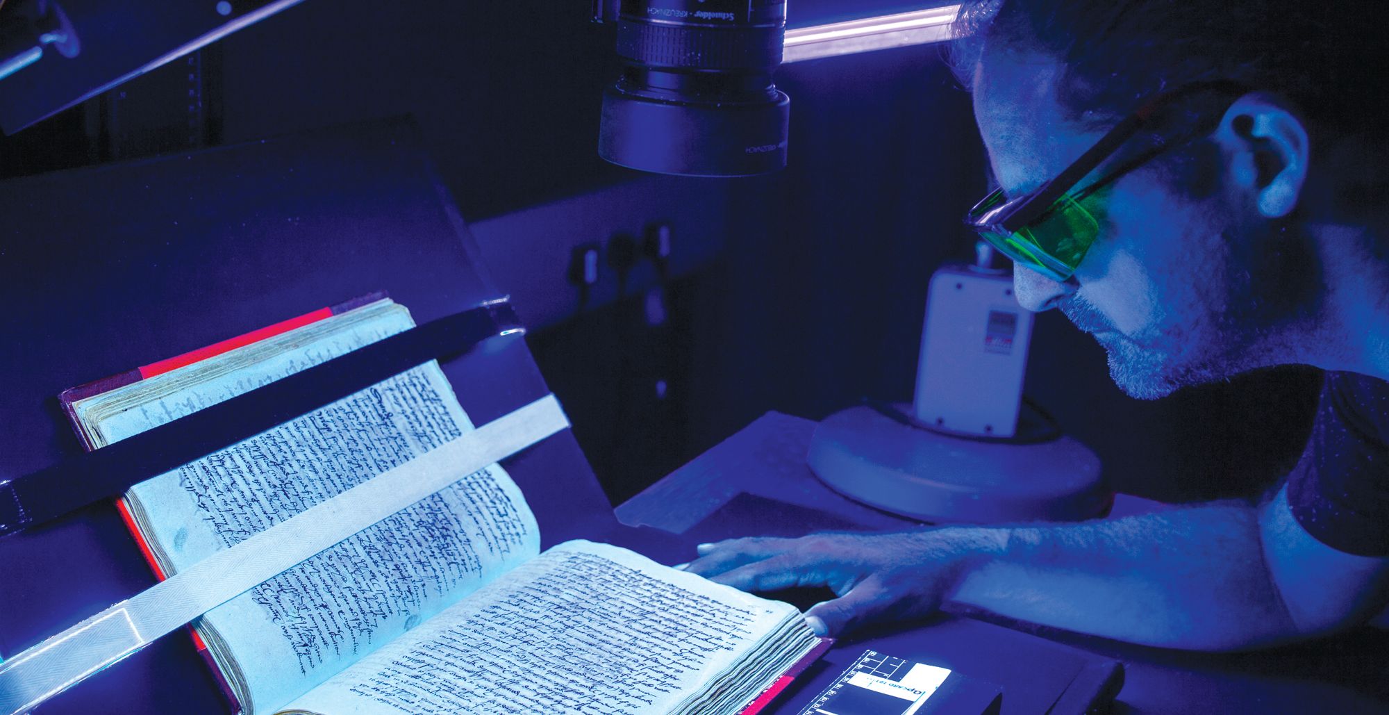 A man wears yellow glasses and looks closely at a book during multi-spectral imaging at the British Library.