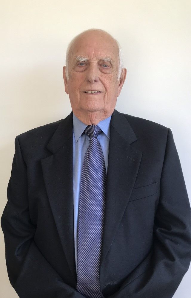 Elderly man in a dark suit, light blue shirt, and patterned tie, standing against a plain white background.